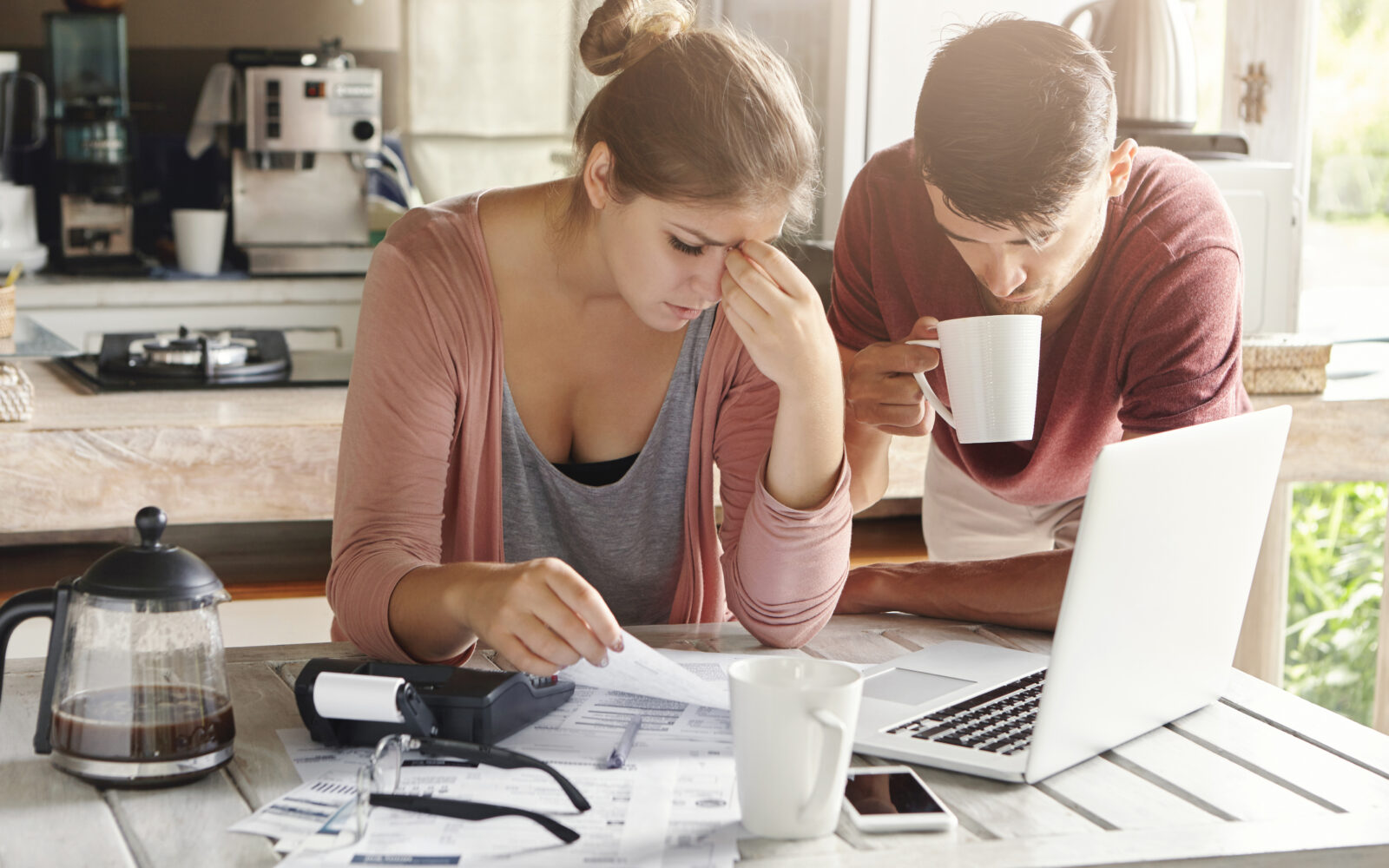 Couple facing financial problem, failing to pay loan in bank. Stressed woman managing family budget, making calculations using laptop and calculator, her husband standing next to her with cup of tea